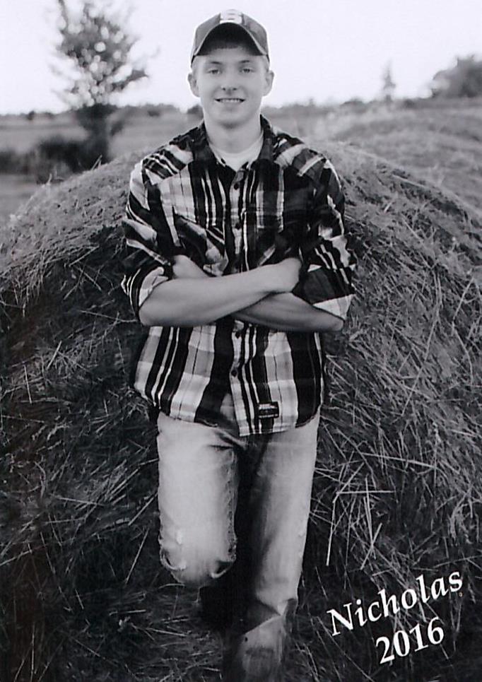 smiling young man with arms crossed leaning up against large bale of hay