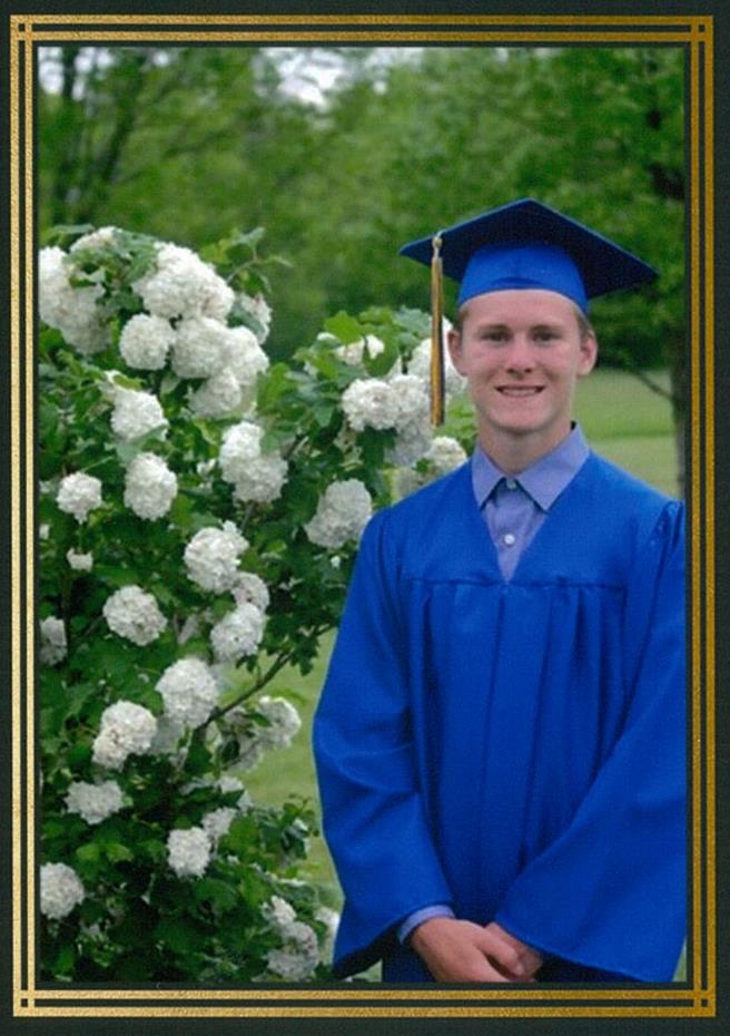 smiling young man in blue cap and gown next to white flowered bush
