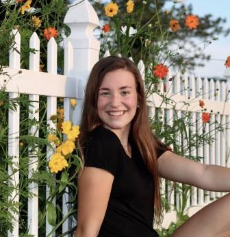 smiling young woman leaning up against white fence with flowers poking through