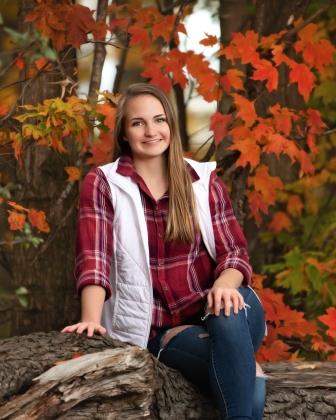 smiling woman in flannel and vest sitting on log in front of fall foliage