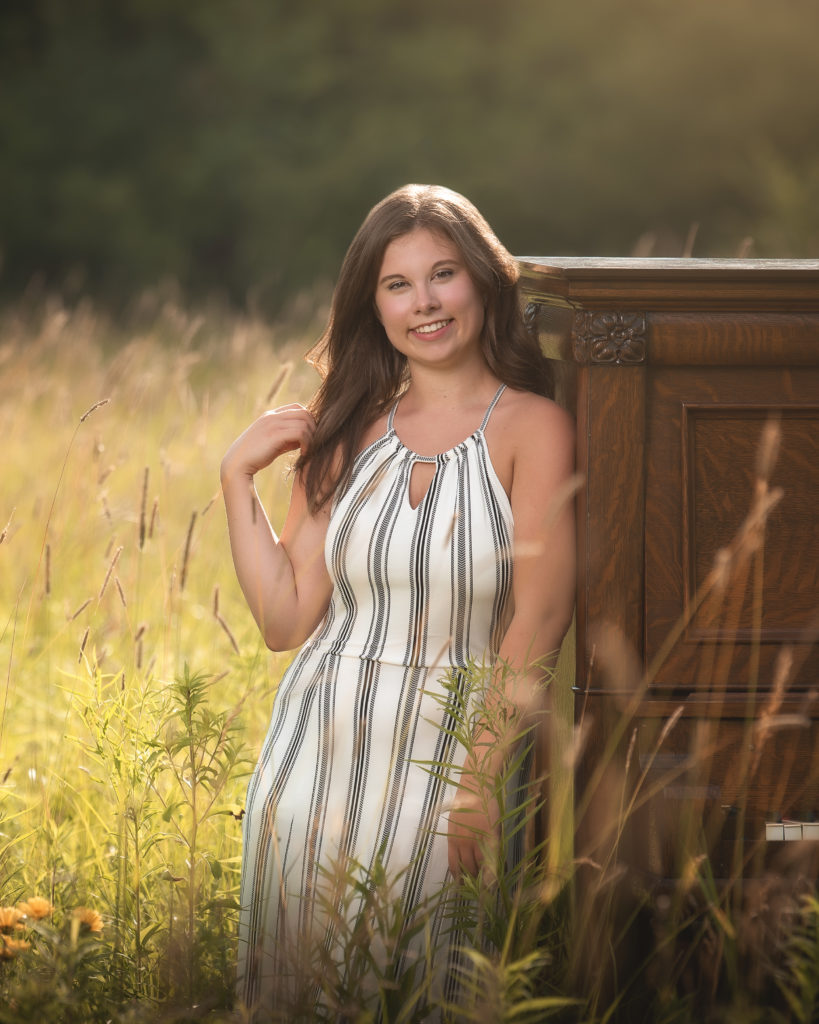 smiling young woman in a field leaning up against a large wooden piano