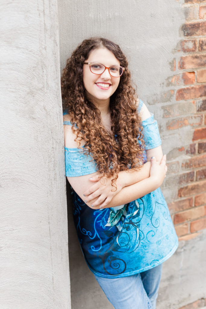 smiling young woman with long curly hair and red glasses leaning up against a wall