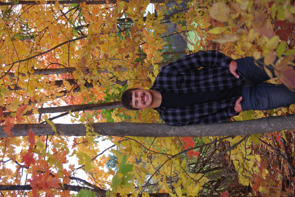 smiling young man with mustache leaning up against a thin tree with colorful leaves