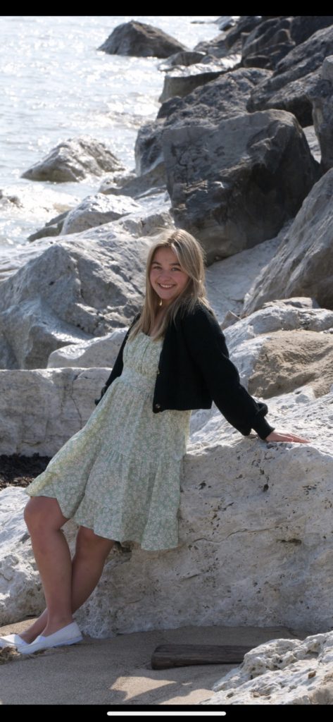 young woman wearing a dress and black cardigan leaning up against a large rock in a harbor