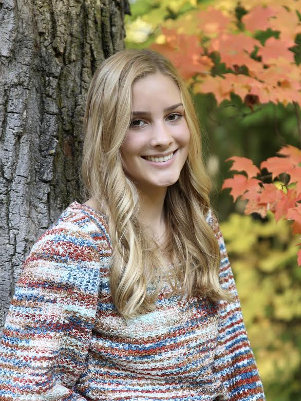 young smiling woman with long blonde hair standing against a tree with colorful leaves