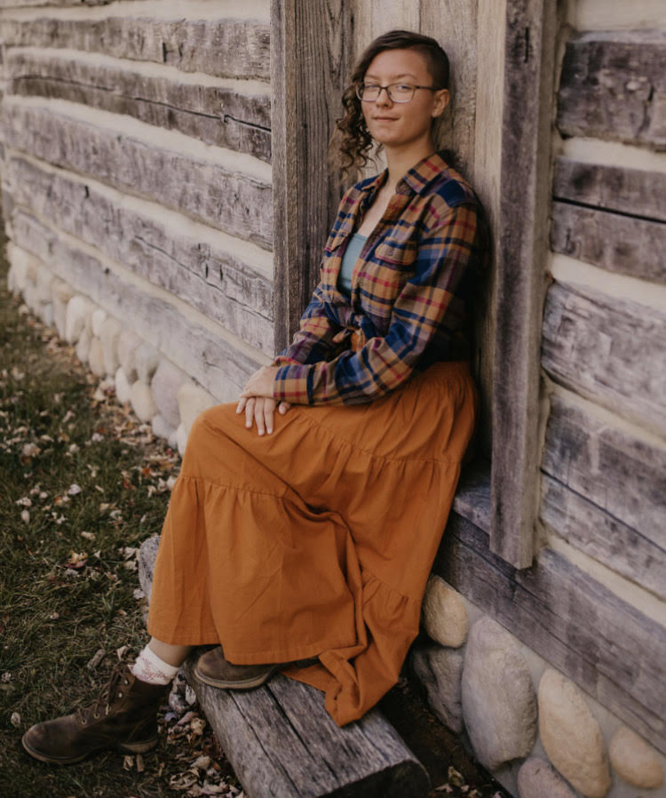 young person with glasses, wearing a plaid shirt and skirt sitting in a doorway of a log house