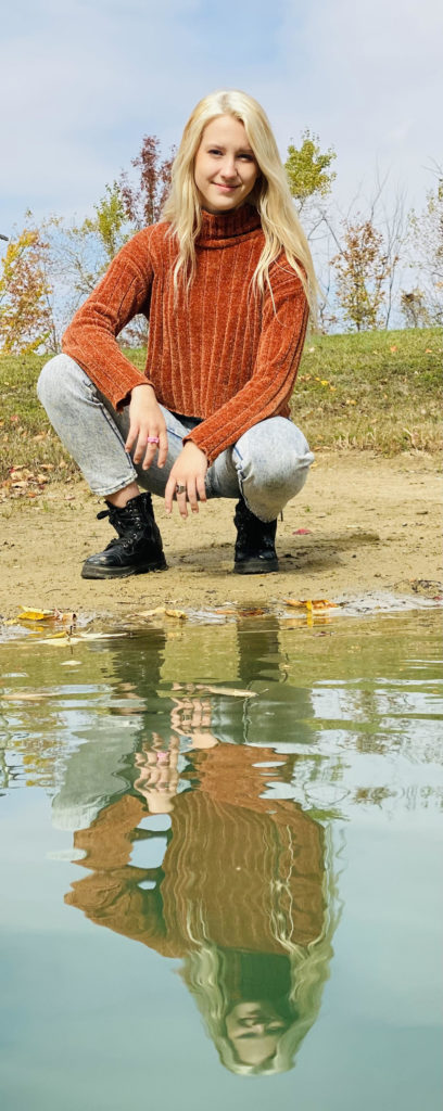 young woman with blonde hair, sweater, and jeans kneeling next to water
